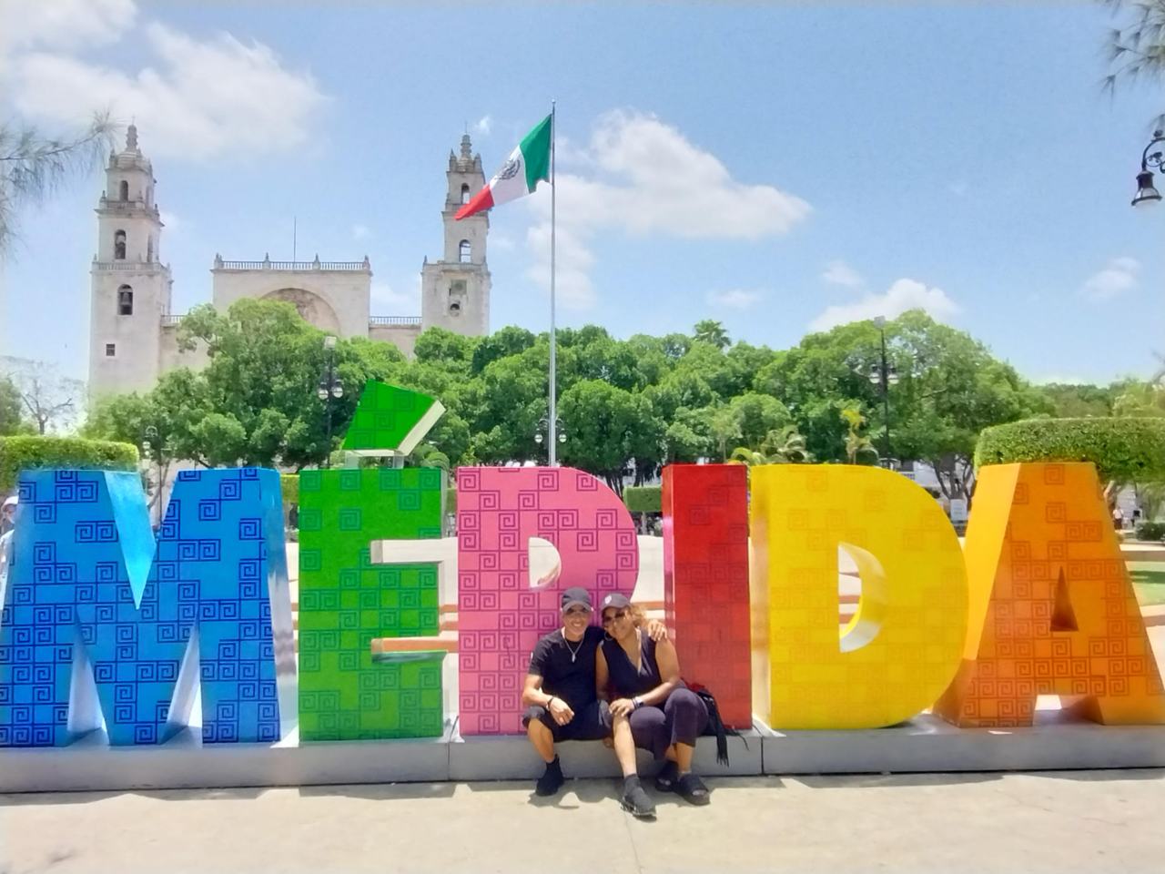 Viajeros sentados en las letras de colores de Mérida frente a la catedral